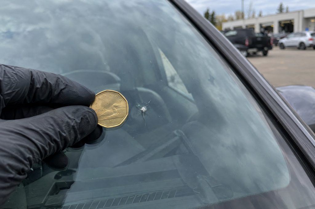 A gloved hand holding a loonie next to a small star-shaped chip in a windshield
